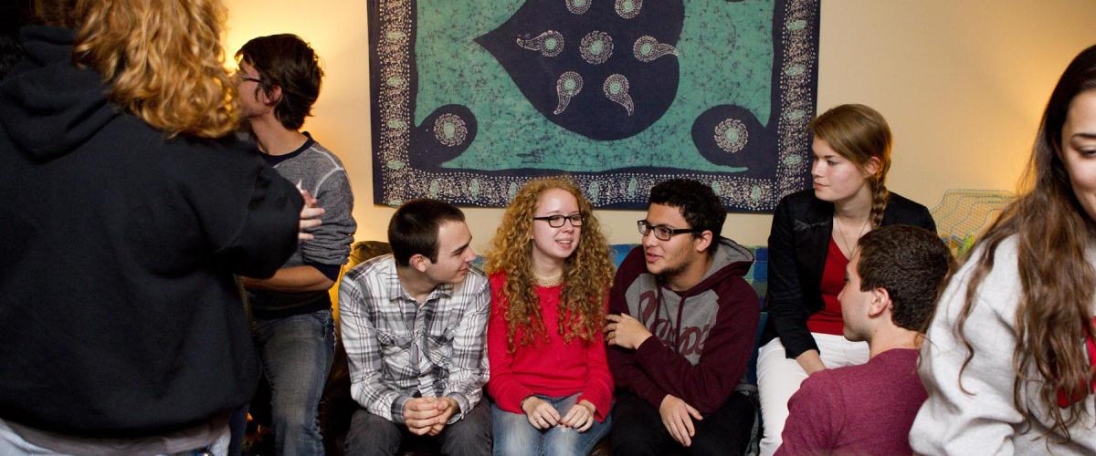 Four students sit together on a couch chatting with one another while other students mingle around them